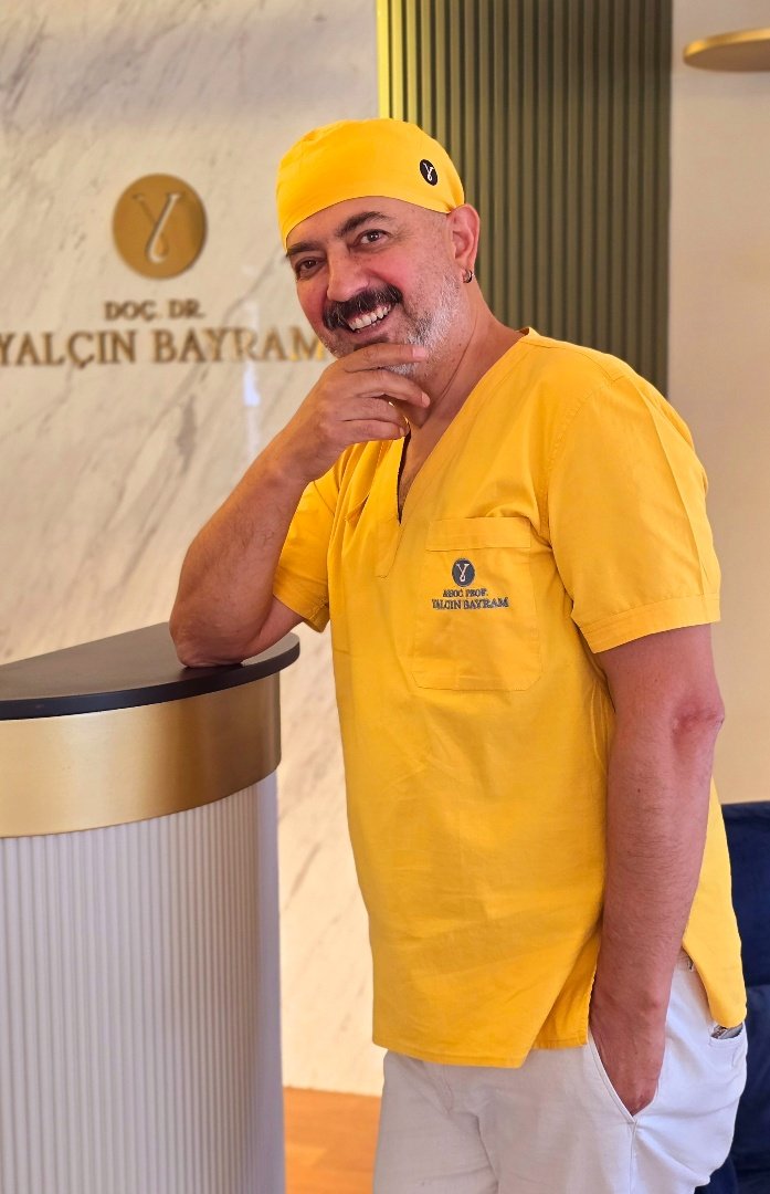 Assoc. Prof. Dr. Yalçın Bayram, Plastic and Aesthetic Surgery Specialist, posing in his clinic wearing yellow surgical scrubs.