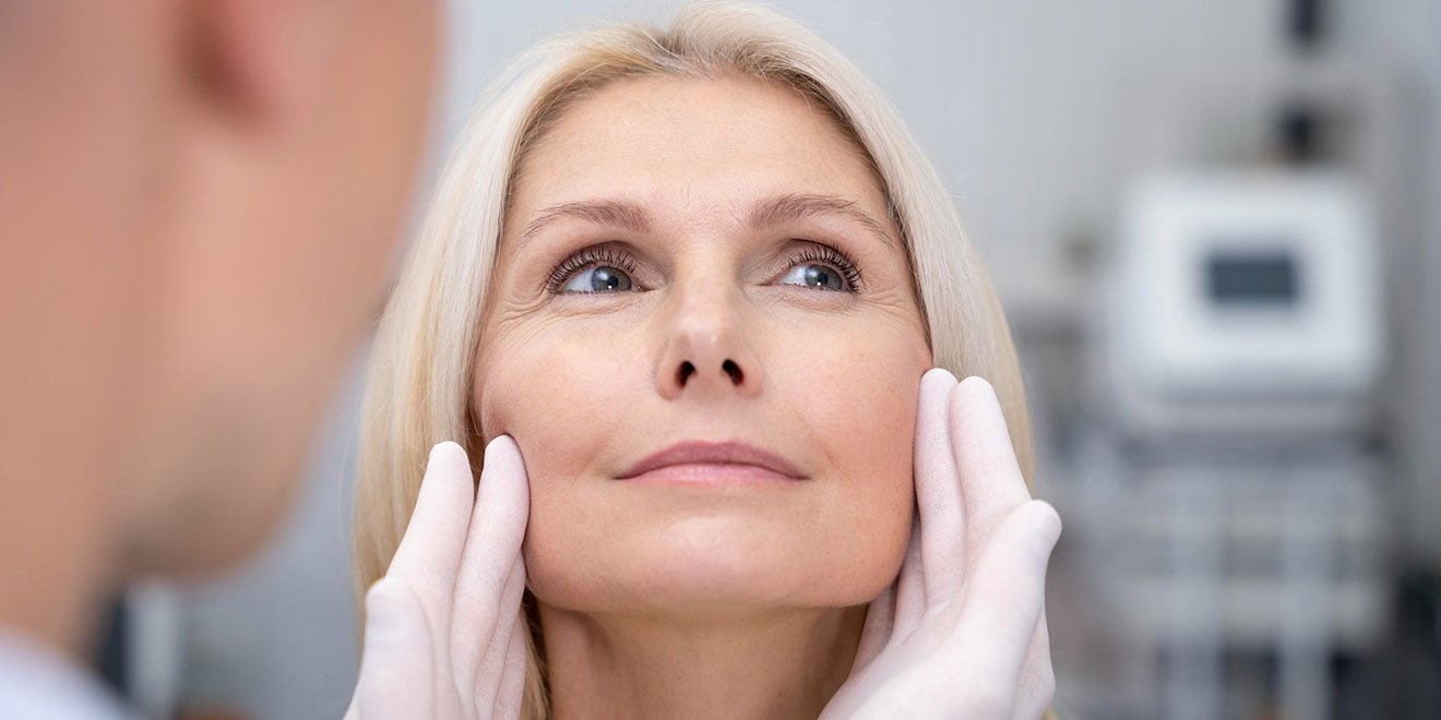 Middle-aged blonde woman having her face examined by doctor during facelift surgery consultation.